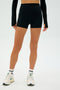 A person in a black long-sleeve top, SPLITS59 Ella Airweight High Waist Short 3.5", white sneakers, and socks stands in a light-colored space.