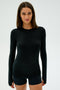 A person with long dark hair, wearing the Louise Rib Long Sleeve - Black by SPLITS59, stands against a plain white background. The black semi-fitted shirt is MADE IN LOS ANGELES and paired with shorts.