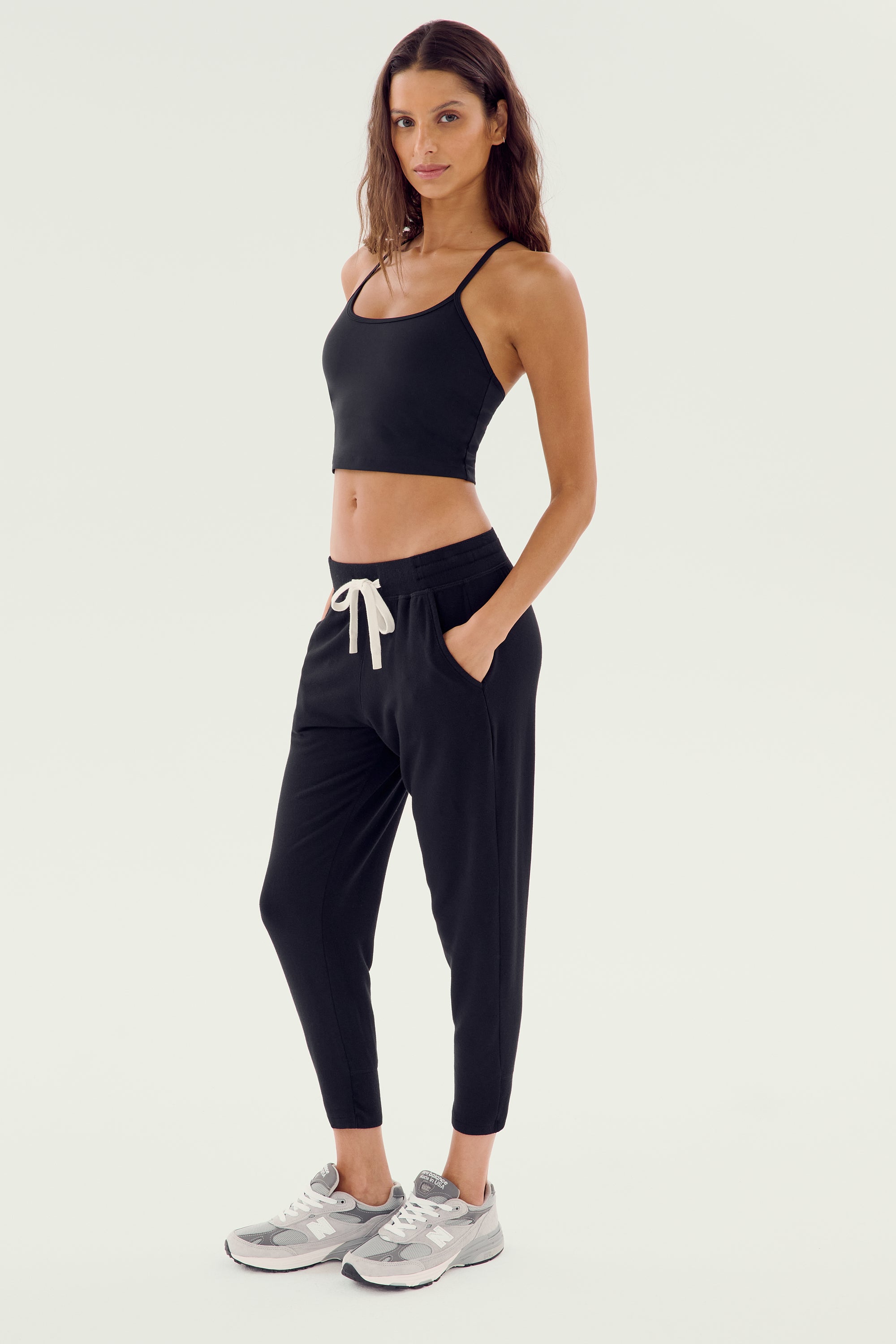 A person stands against a plain background wearing a black crop top, Splits59 Reena Fleece 7/8 Sweatpants - Black with a white drawstring, and gray sneakers—ideal for everyday wear.