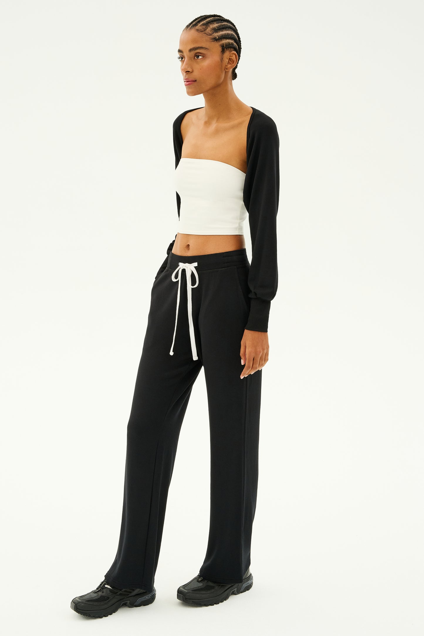 Dressed in a white tube top, black cardigan, and SPLITS59's Raven Fleece Full Length Sweatpant in black paired with sneakers, this individual's style embodies the MADE IN LOS ANGELES aesthetic. Set against a white backdrop, their outfit seamlessly blends the serene ambiance of a luxury spa with the plush comfort of ultra-luxe fleece.