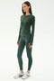 A woman poses in a studio wearing the SPLITS59 Airweight Lite LS Waist Length top in Military, paired with matching leggings and white athletic shoes, set against a plain light background.