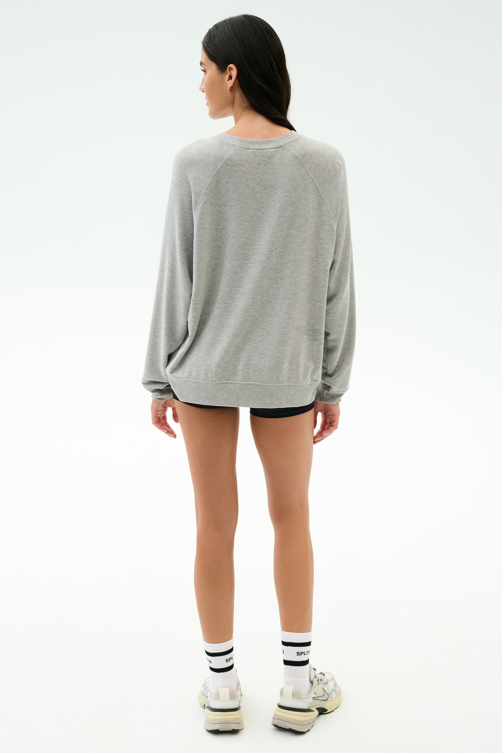Someone stands facing away in the SPLITS59 Andie Oversized Fleece Varsity Graphic Sweatshirt in Heather Grey, paired with black shorts, white socks, and sneakers against a plain light background.