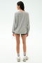 Someone stands facing away in the SPLITS59 Andie Oversized Fleece Varsity Graphic Sweatshirt in Heather Grey, paired with black shorts, white socks, and sneakers against a plain light background.