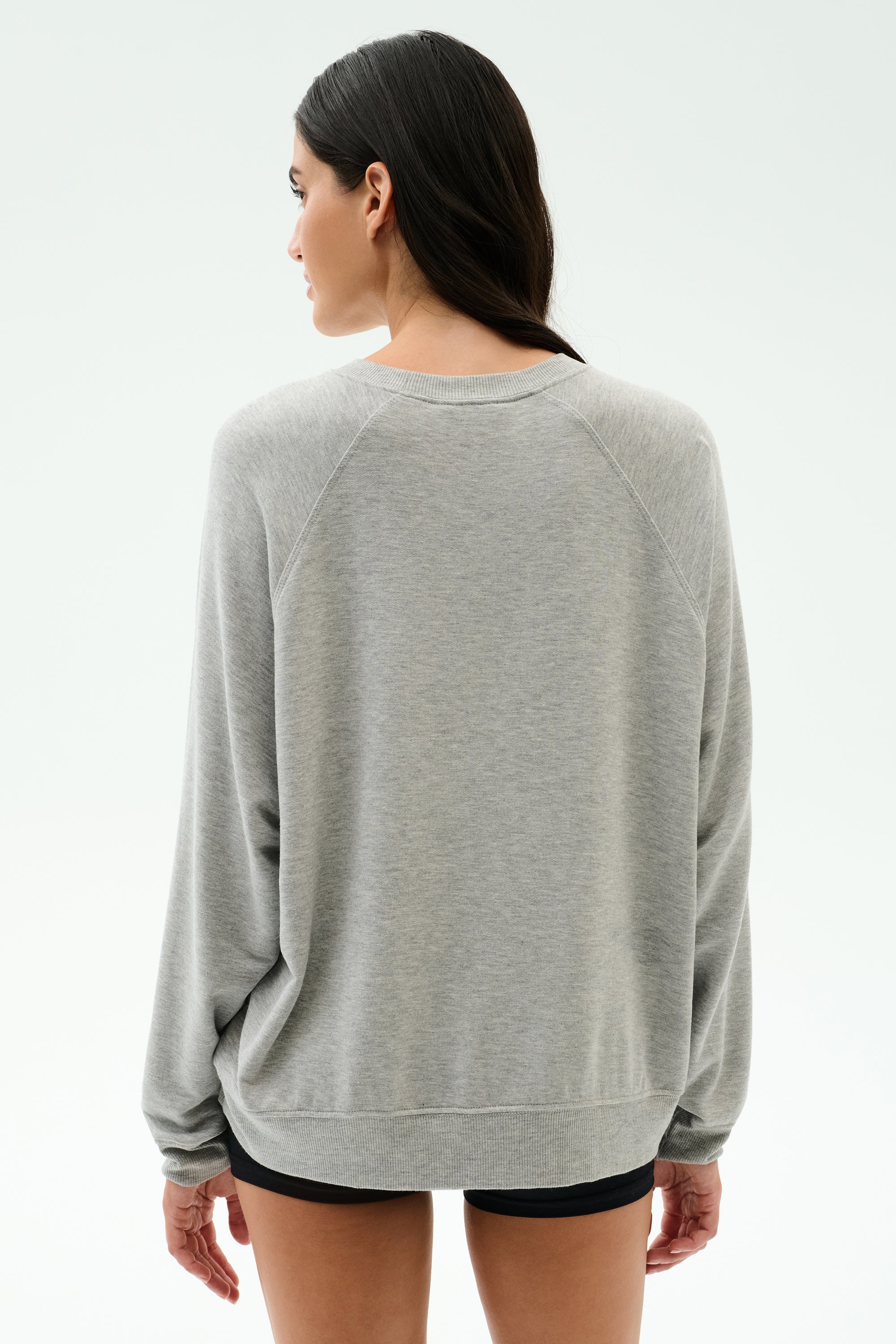 A woman with long dark hair wears the SPLITS59 Andie Oversized Fleece Varsity Graphic Sweatshirt in Heather Grey and black shorts, facing away from the camera against a plain light background.