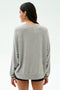 A woman with long dark hair wears the SPLITS59 Andie Oversized Fleece Varsity Graphic Sweatshirt in Heather Grey and black shorts, facing away from the camera against a plain light background.