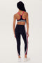 A woman with braided hair wearing a SPLITS59 Bianca Rigor Bra - Black and black nylon-spandex leggings with white and red stripes stands facing away from the camera. She is also wearing white sneakers.