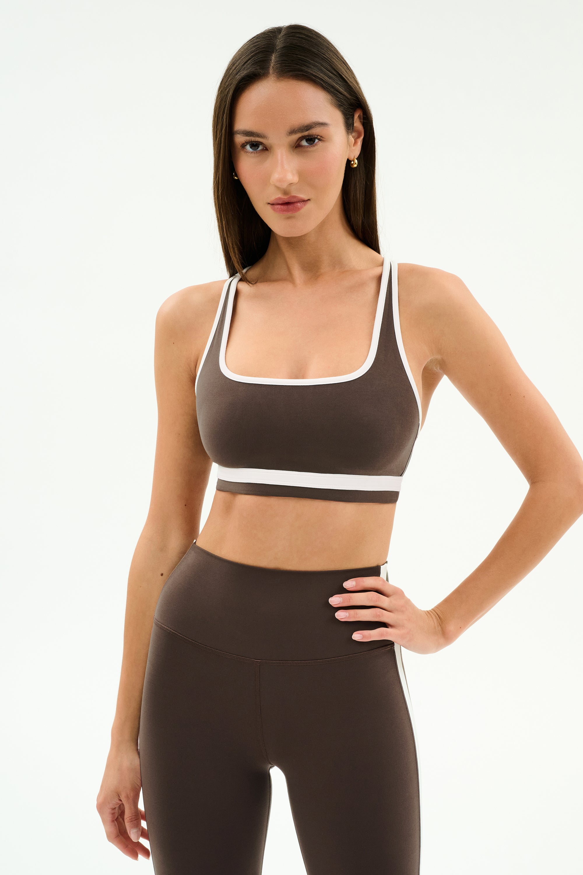 A woman stands against a plain white background wearing the SPLITS59 Bianca Rigor Bra in Dark Chocolate/White with matching high-waisted leggings, an ideal set for high-impact workouts.