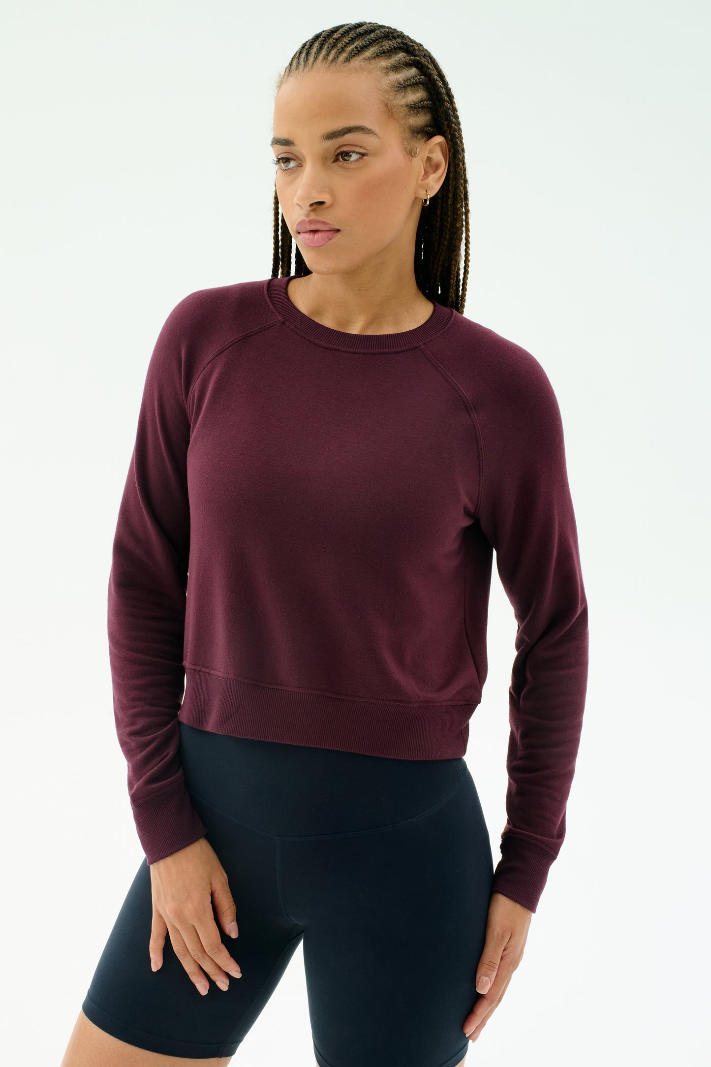 A person with braided hair wears the SPLITS59 Brooks Fleece Long Sleeve Sweatshirt in Dark Cherry and black shorts, standing against a plain white background.