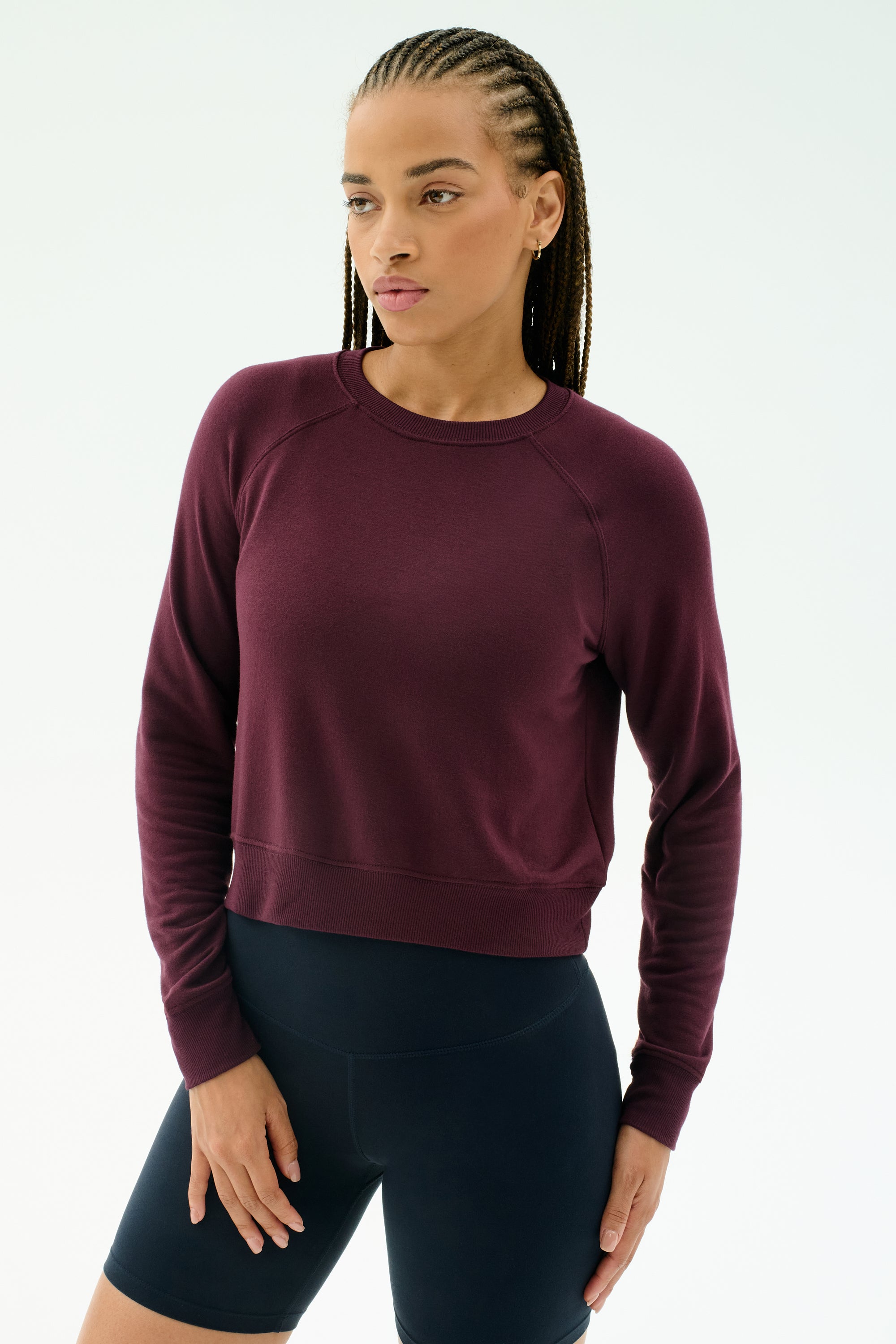 A person with braided hair wears the SPLITS59 Brooks Fleece Long Sleeve Sweatshirt in Dark Cherry and black shorts, standing against a plain white background.