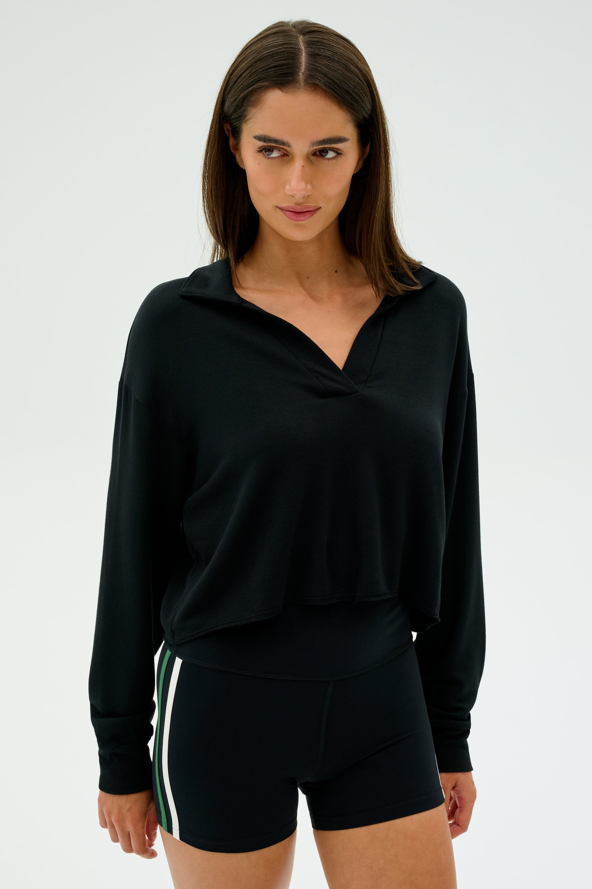 A person in a black SPLITS59 Joan Fleece Polo Sweatshirt and black shorts with green and white stripes stands against a plain background.