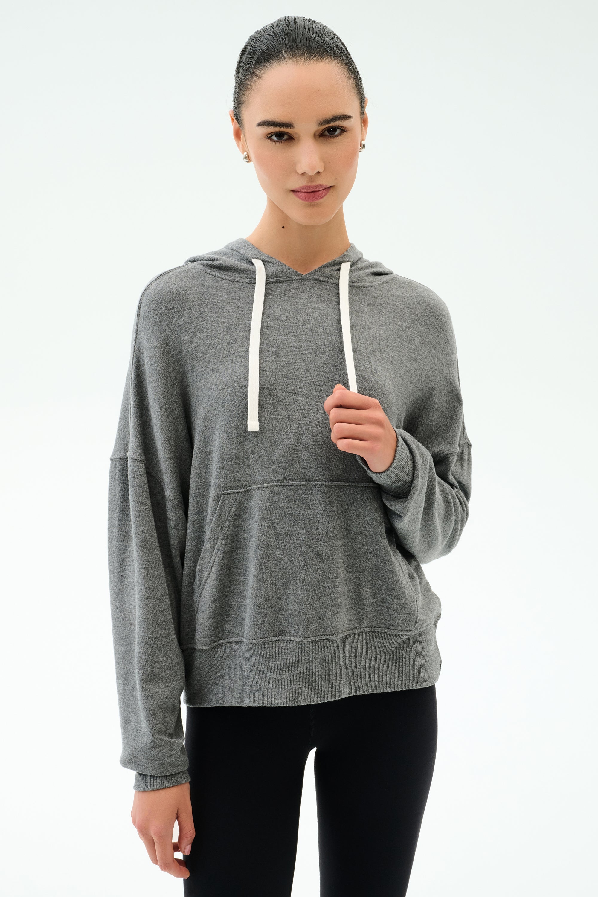 A woman wears the SPLITS59 Joey Long Sleeve Hoodie in Medium Heather Grey and black spandex leggings, standing against a plain light background.