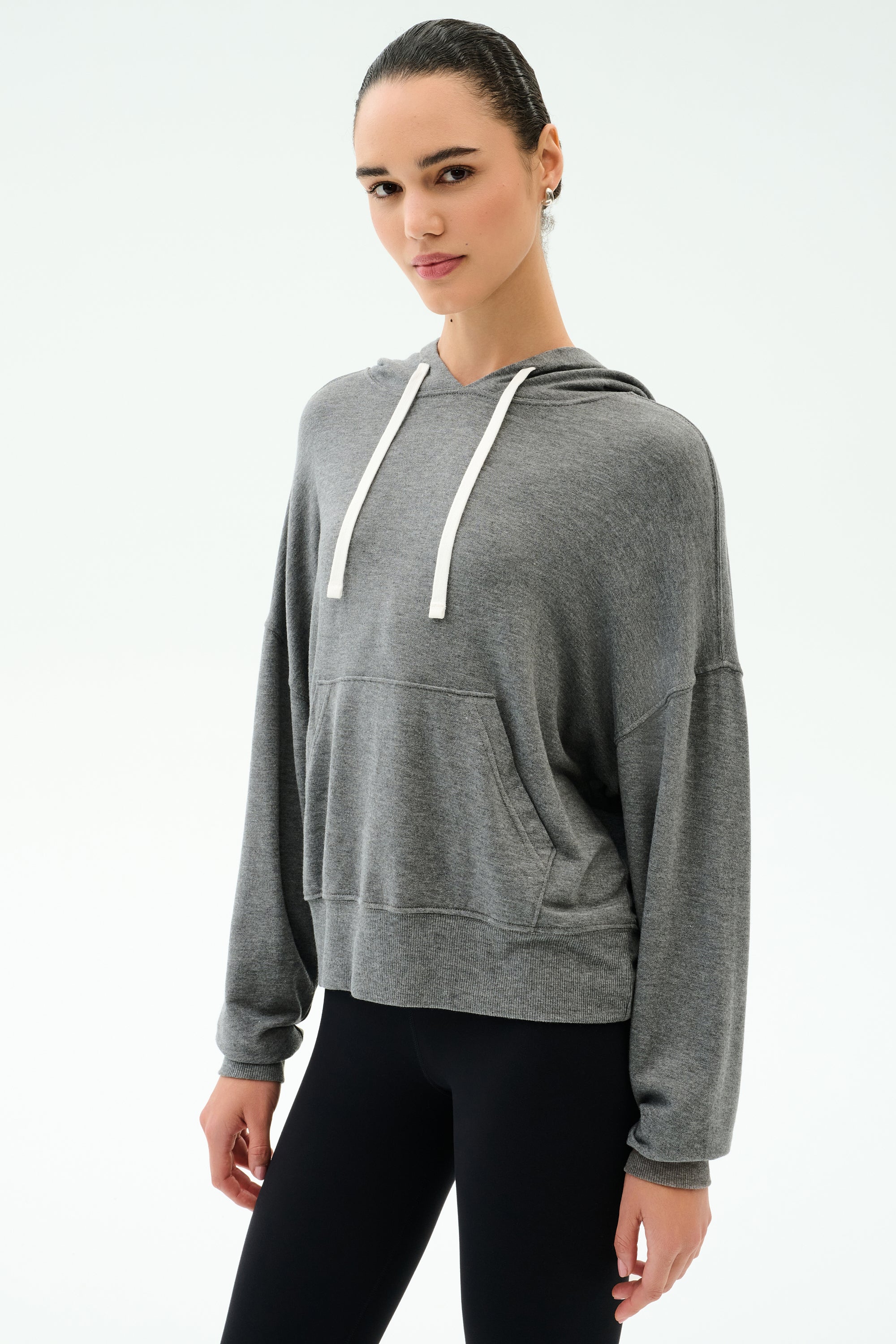A person wears the SPLITS59 Joey Long Sleeve Hoodie in Medium Heather Grey and black spandex leggings, standing against a plain light background.