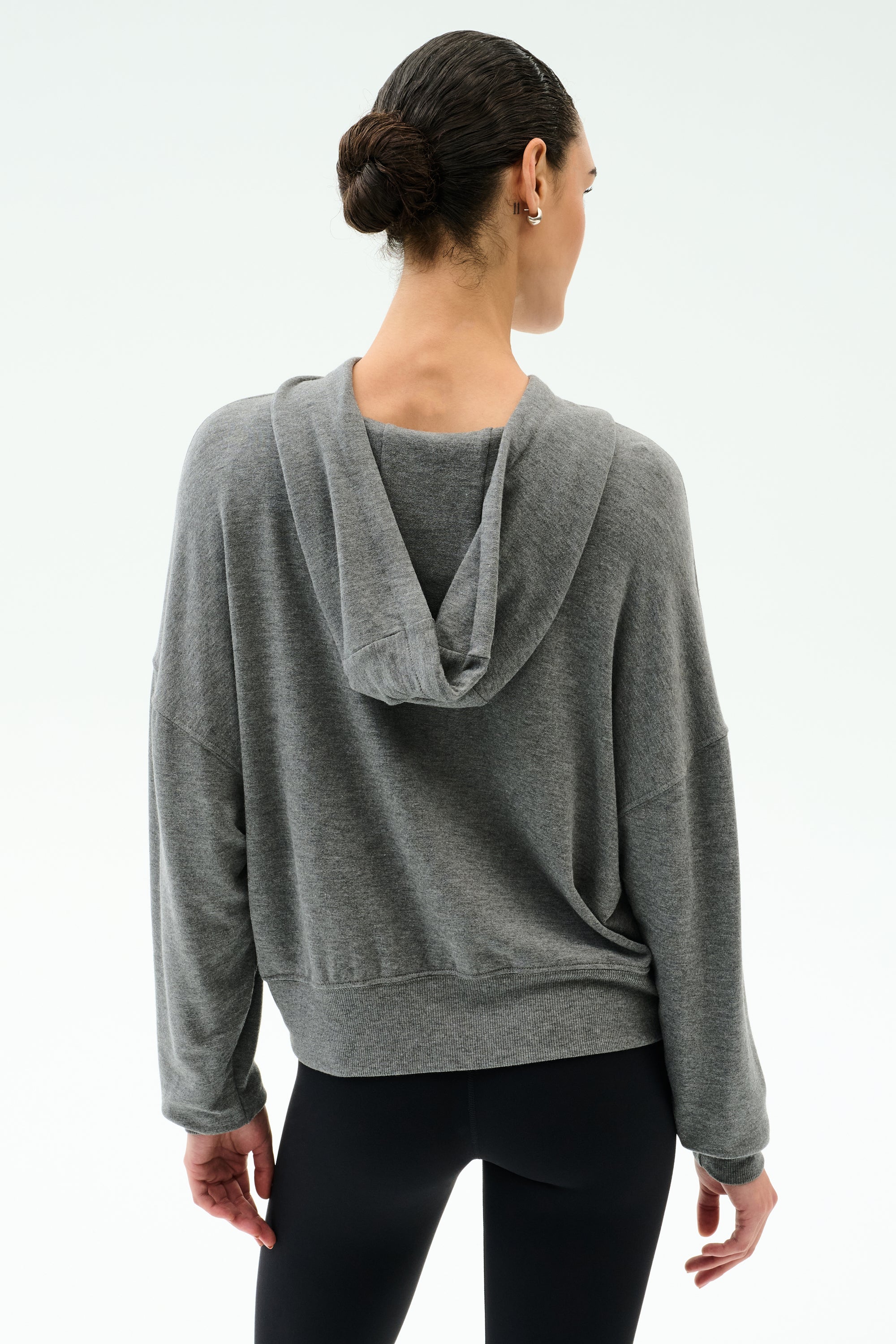A person is seen from behind wearing the SPLITS59 Joey Long Sleeve Hoodie in Medium Heather Grey and black spandex leggings, with hair in a neat bun, set against a plain white background.