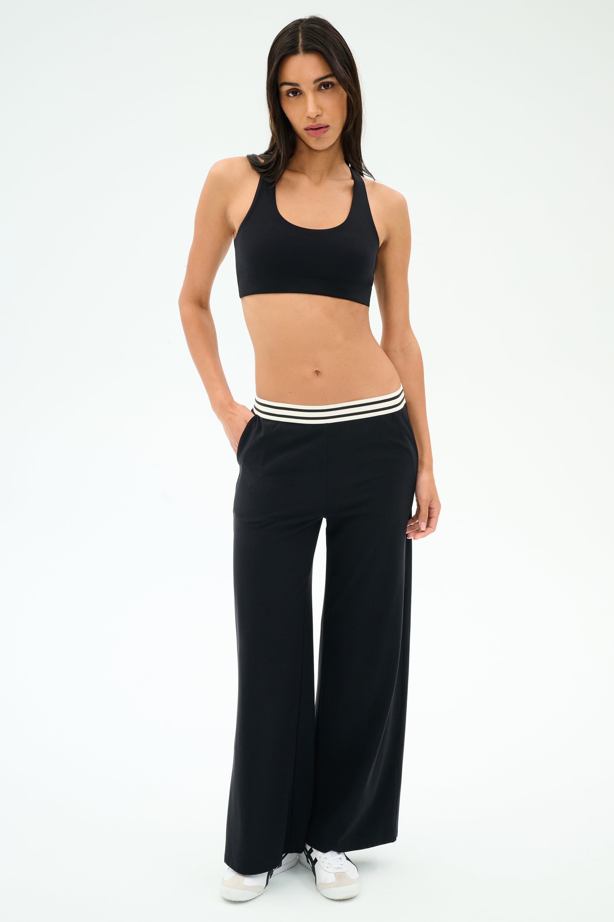 A woman stands against a plain background, wearing the SPLITS59 Michele Airweight Trouser in black, paired with a black sports bra and white sneakers.