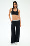 A woman stands against a plain background, wearing the SPLITS59 Michele Airweight Trouser in black, paired with a black sports bra and white sneakers.