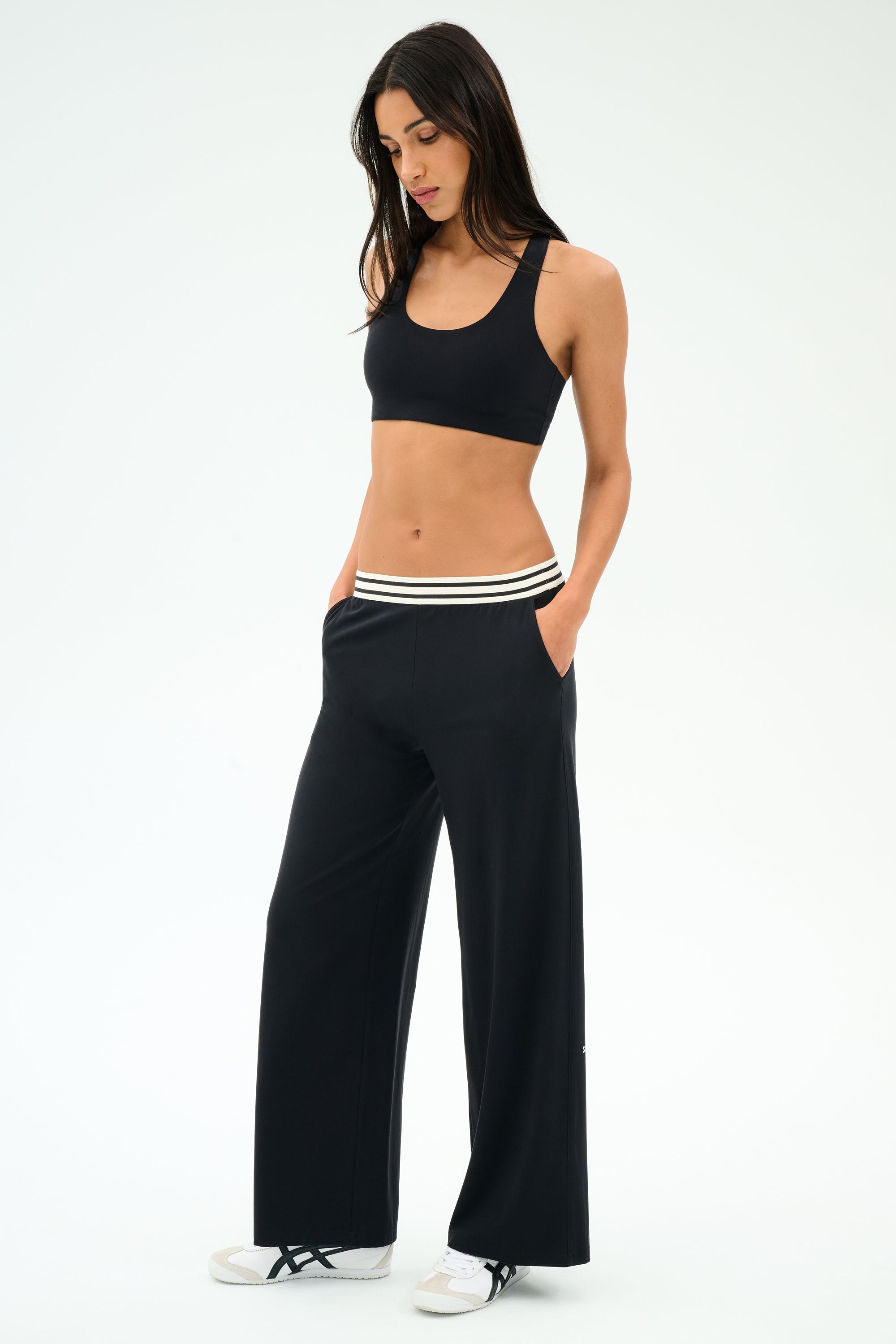 A woman stands with hands in pockets, wearing SPLITS59 Michele Airweight Trouser in black, a black sports bra, and white sneakers against a plain light background.