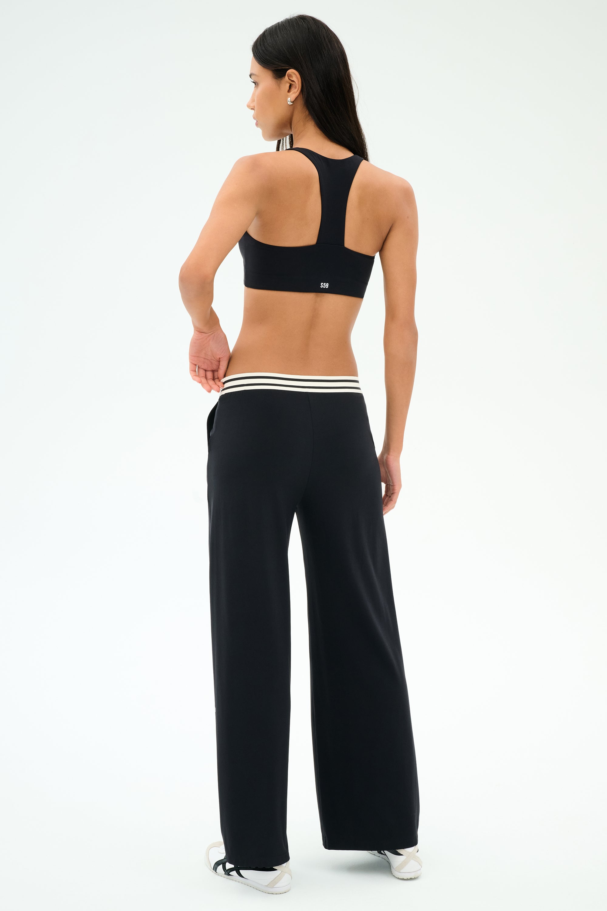 A woman faces away, wearing SPLITS59 Michele Airweight Trouser in black with white-striped waistband, a black sports bra, and white sneakers against a plain background.