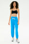 A person stands facing forward, wearing a black modal crop top, SPLITS59's Andie Oversized Fleece Sweatpant in Riviera blue with a white drawstring, and silver spandex athletic shoes against a plain white background.
