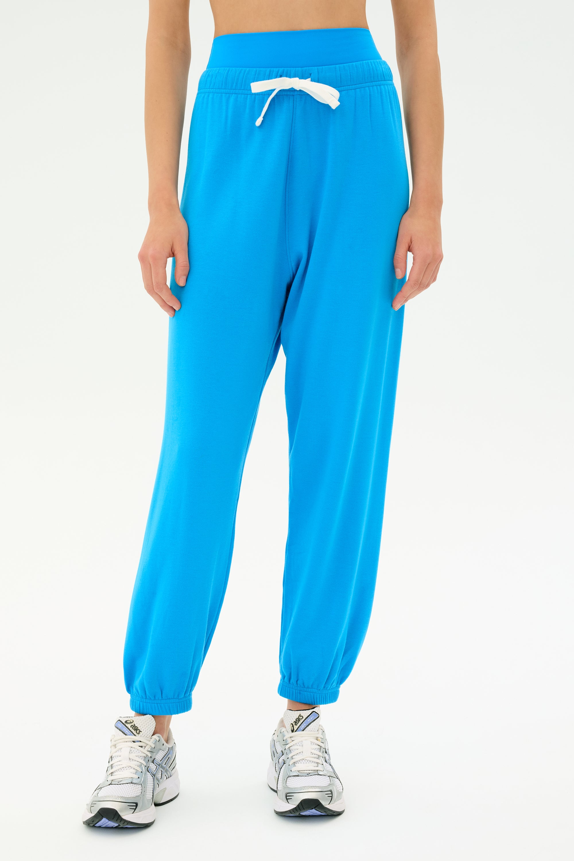 Model wears the SPLITS59 Andie Oversized Fleece Sweatpant in Riviera, a bright blue pant with white drawstring, paired with silver sneakers and set against a plain white background.