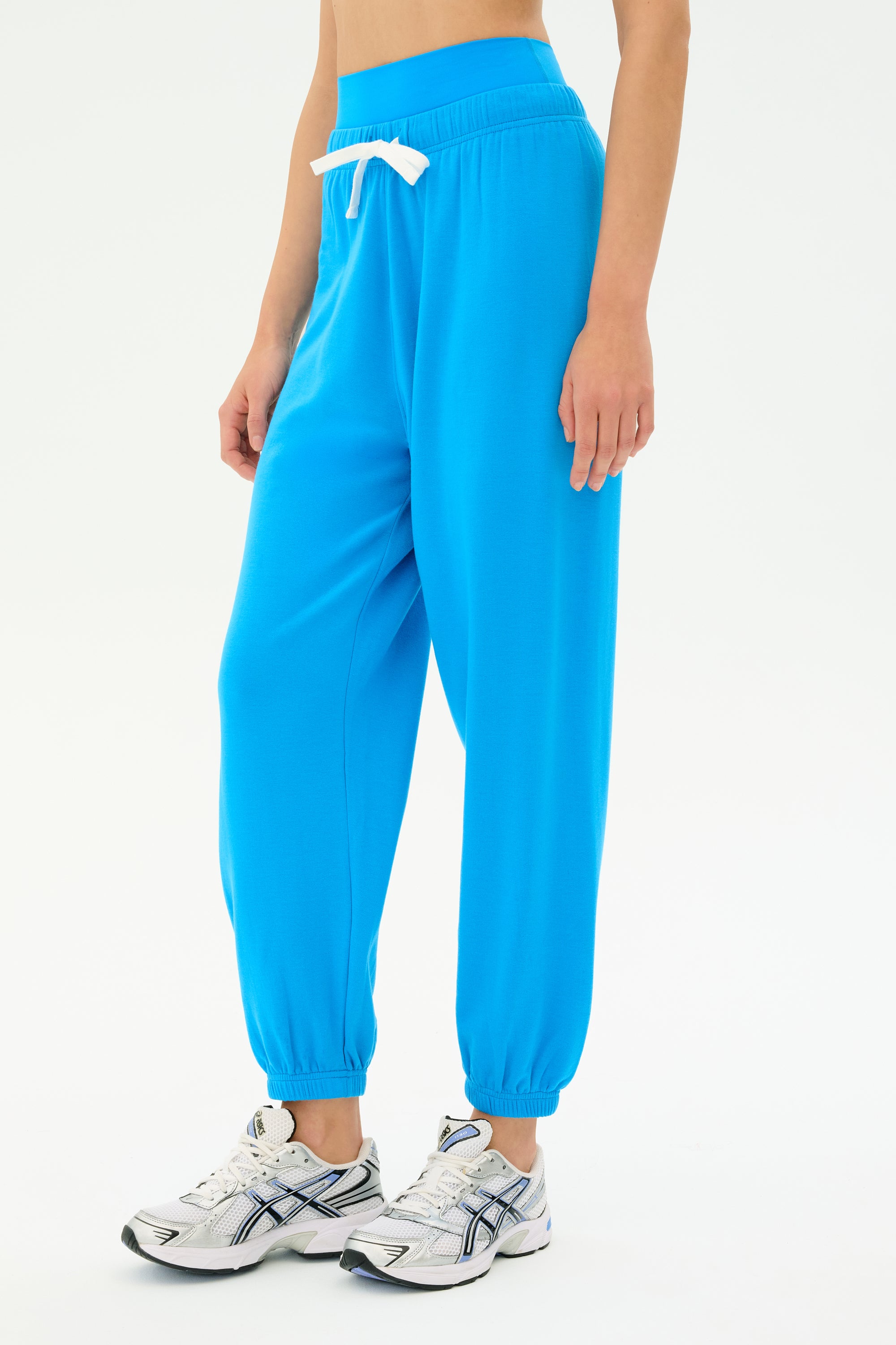 A person wears SPLITS59's Andie Oversized Fleece Sweatpant in Riviera, bright blue with a white drawstring, paired with white athletic sneakers, and stands against a plain light background.