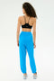 A woman with curly hair faces away, wearing a black spandex sports bra, white sneakers, and SPLITS59 Andie Oversized Fleece Sweatpant in Riviera blue against a plain light background.