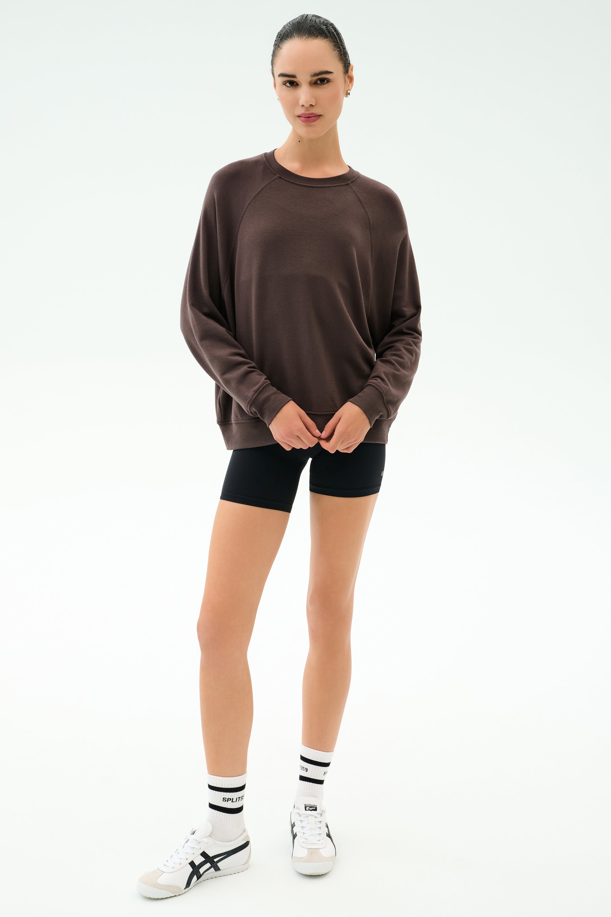 A woman stands facing forward against a white background, wearing the SPLITS59 Andie Oversized Fleece Sweatshirt in Dark Chocolate with black shorts, white socks with black stripes, and white sneakers.