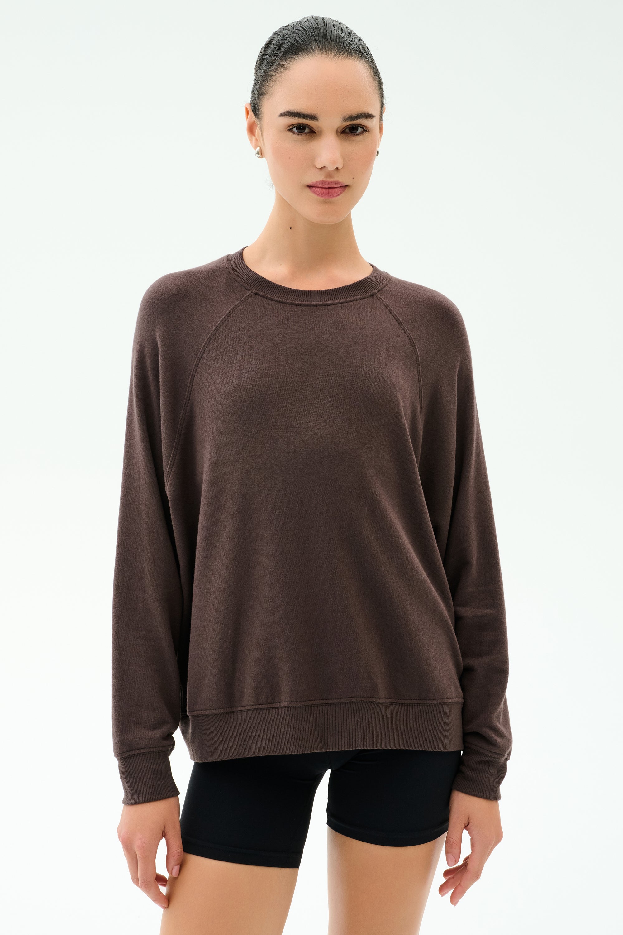 A woman wears the SPLITS59 Andie Oversized Fleece Sweatshirt in Dark Chocolate with black shorts, standing against a plain white background.