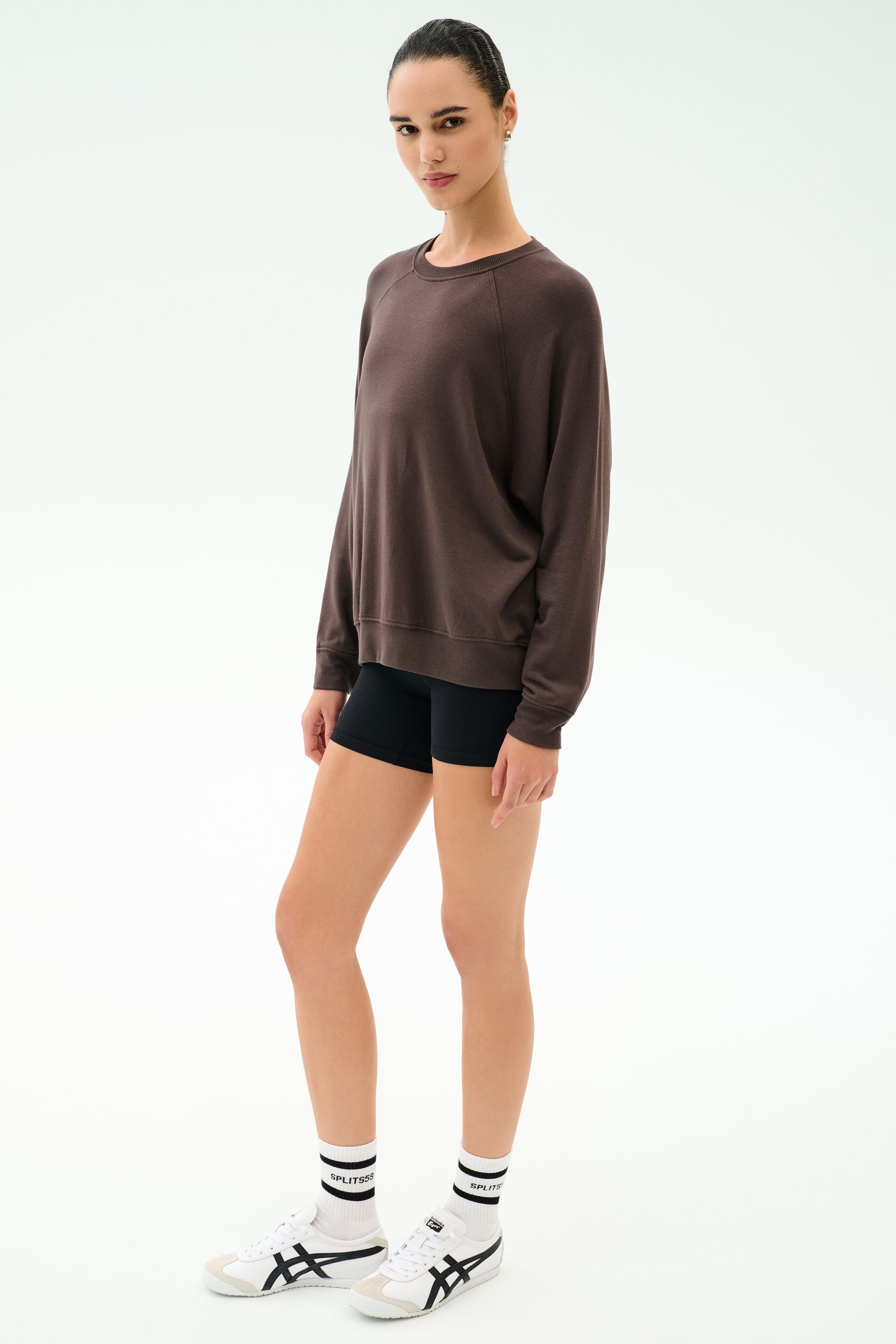 A person stands against a plain background wearing the SPLITS59 Andie Oversized Fleece Sweatshirt in Dark Chocolate, paired with black shorts, white socks, and white sneakers.