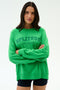 A woman with long dark hair wears the SPLITS59 Andie Oversized Fleece Varsity Graphic Sweatshirt in Grass and black shorts, standing against a plain white background with arms crossed.