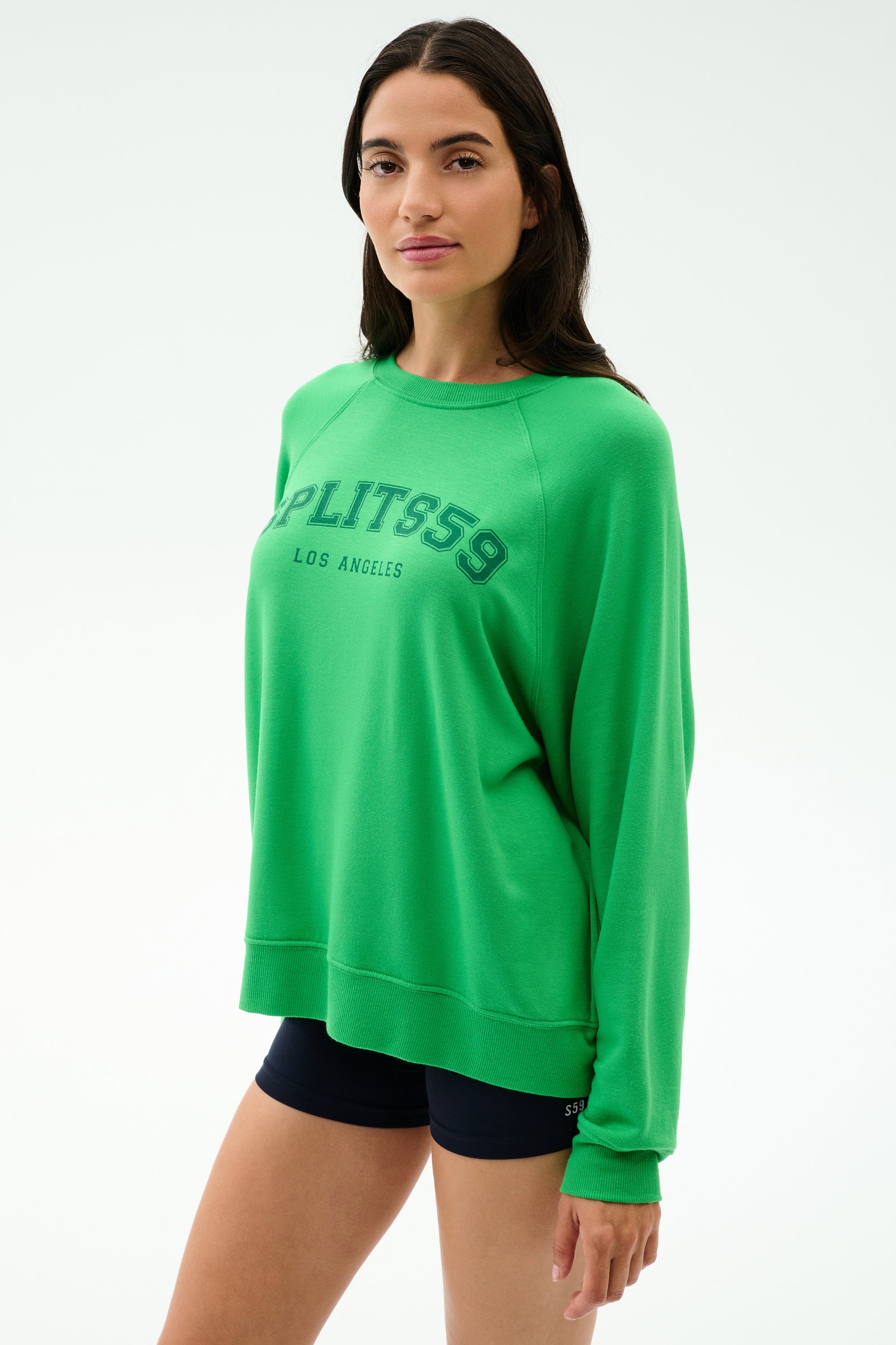 A woman wears the SPLITS59 Andie Oversized Fleece Varsity Graphic Sweatshirt in Grass/Splits59 Los Angeles, paired with black shorts, standing against a plain light background.
