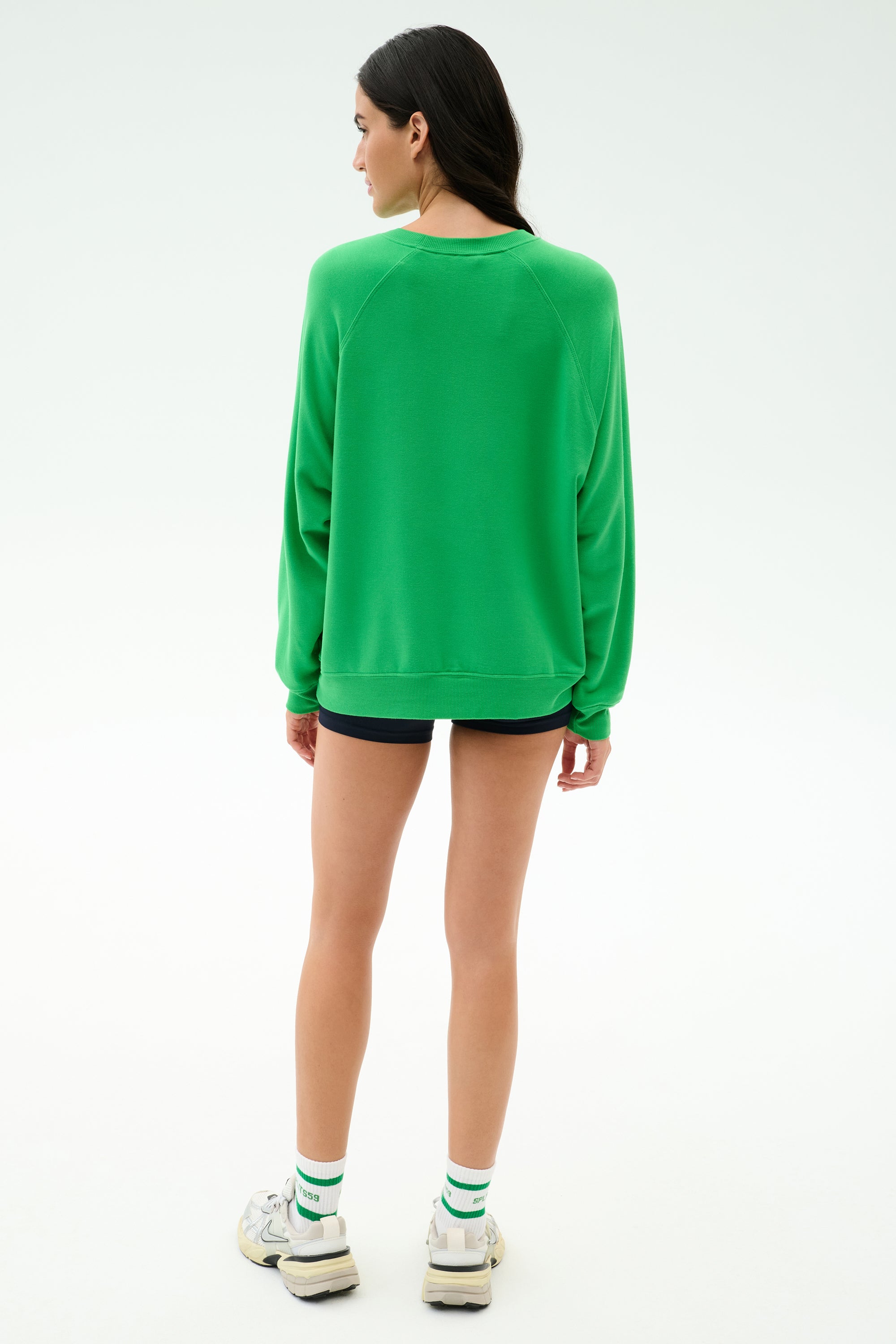 A person stands facing away in the SPLITS59 Andie Oversized Fleece Varsity Graphic Sweatshirt in Grass with black shorts, white socks, and sneakers against a plain light background. This cozy crewneck pullover is made in Los Angeles.