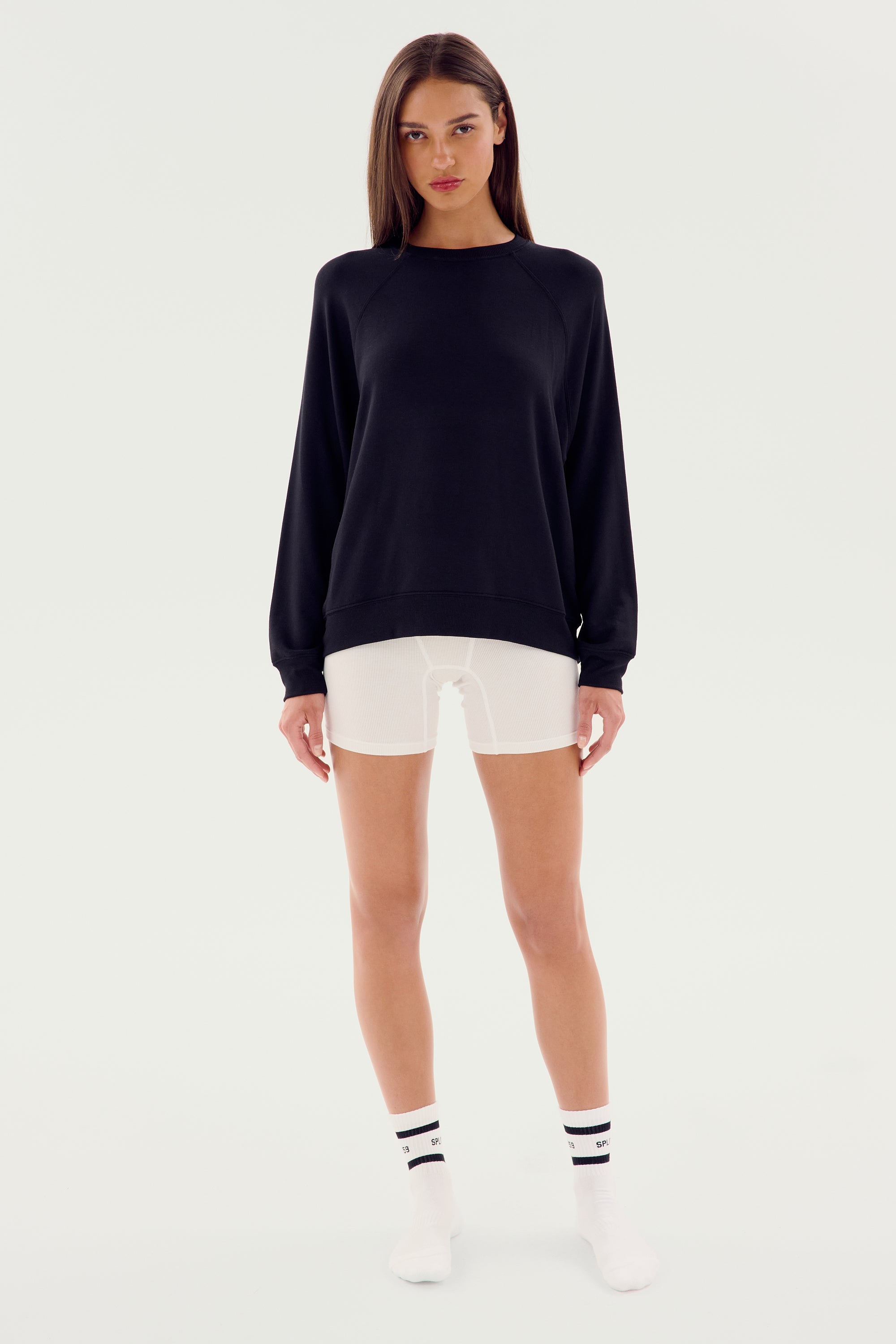 A person standing against a plain background, wearing an SPLITS59 Andie Fleece Sweatshirt - Black made in Los Angeles from stretch modal fabric, white shorts, white socks with black stripes, and white shoes.