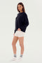 A person with long hair stands sideways, wearing a SPLITS59 Andie Fleece Sweatshirt - Black, white shorts, and white socks with black stripes against a plain light background.