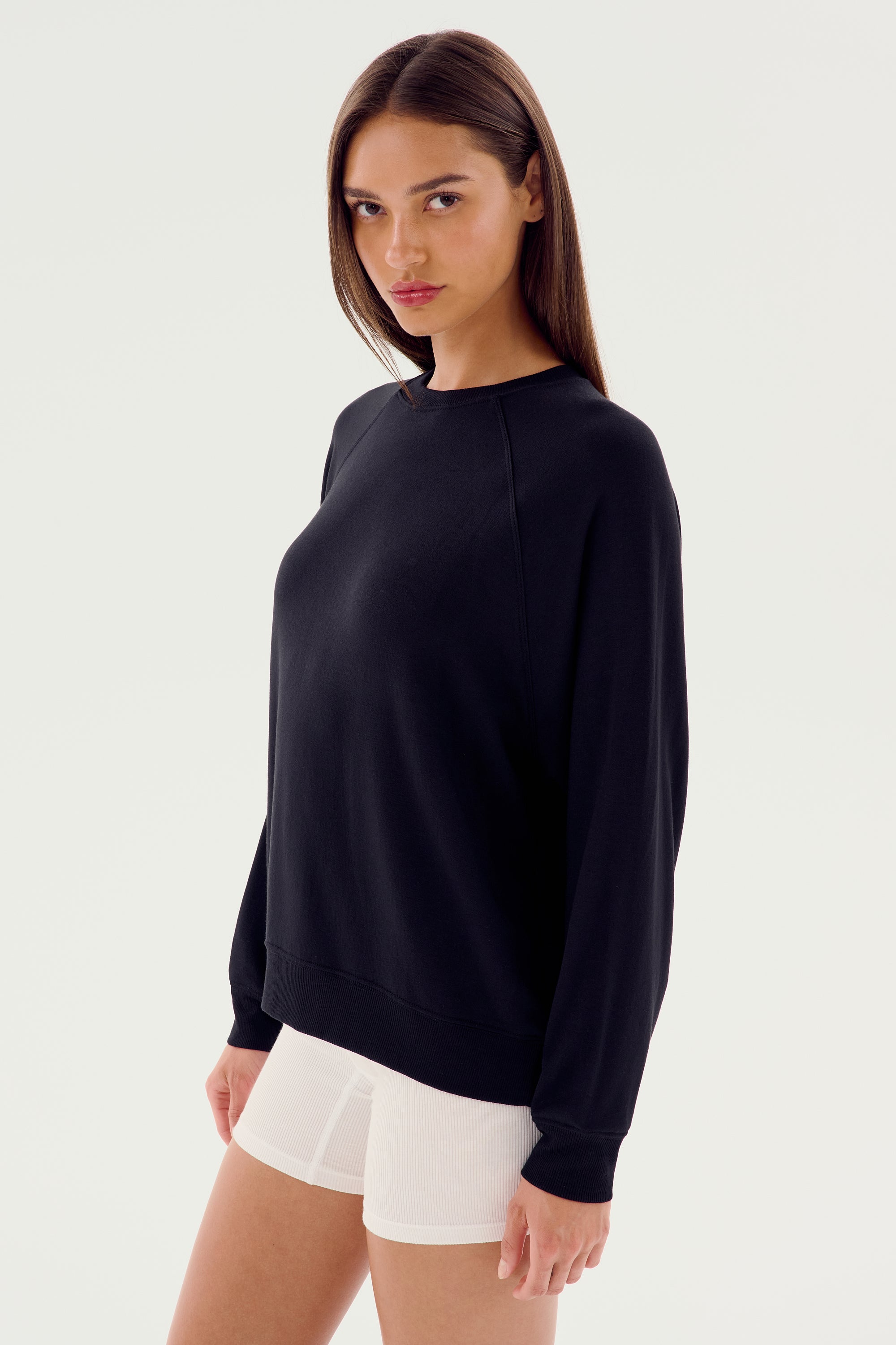 A person stands wearing the SPLITS59 Andie Fleece Sweatshirt - Black and white shorts, facing the camera with a neutral expression. The background is plain and white.