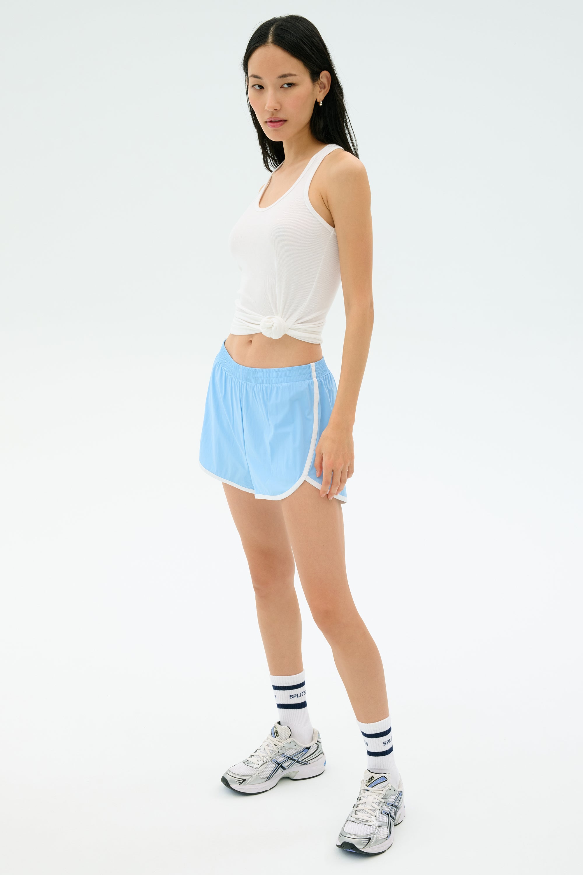 A woman stands against a plain white background wearing the SPLITS59 Megan Woven Nylon Run Short in Skylight, a white spandex tank top, striped socks, and sneakers.