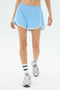 A person wearing SPLITS59 Megan Woven Nylon Run Shorts in Skylight, white socks with blue stripes, and silver running shoes walks against a plain white background.