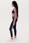 A person with long braided hair stands in profile wearing a dark SPLITS59 Bianca Rigor Bra - Indigo, matching high-waisted leggings with green and white stripes made of spandex, and white athletic shoes.