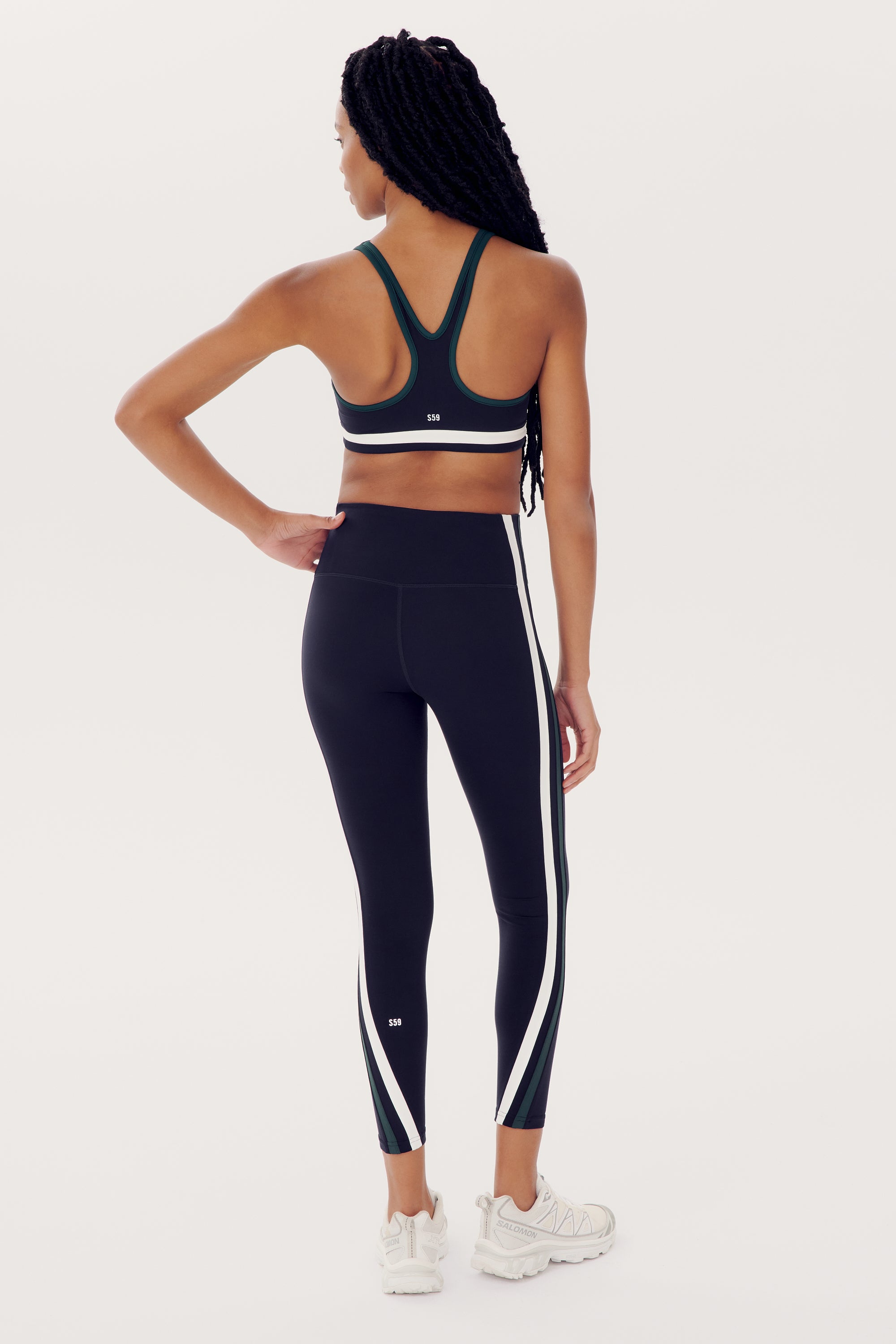 A woman stands facing away, wearing a SPLITS59 Bianca Rigor Bra - Indigo, black nylon athletic leggings with white side stripes, and white sneakers. She has long braided hair.