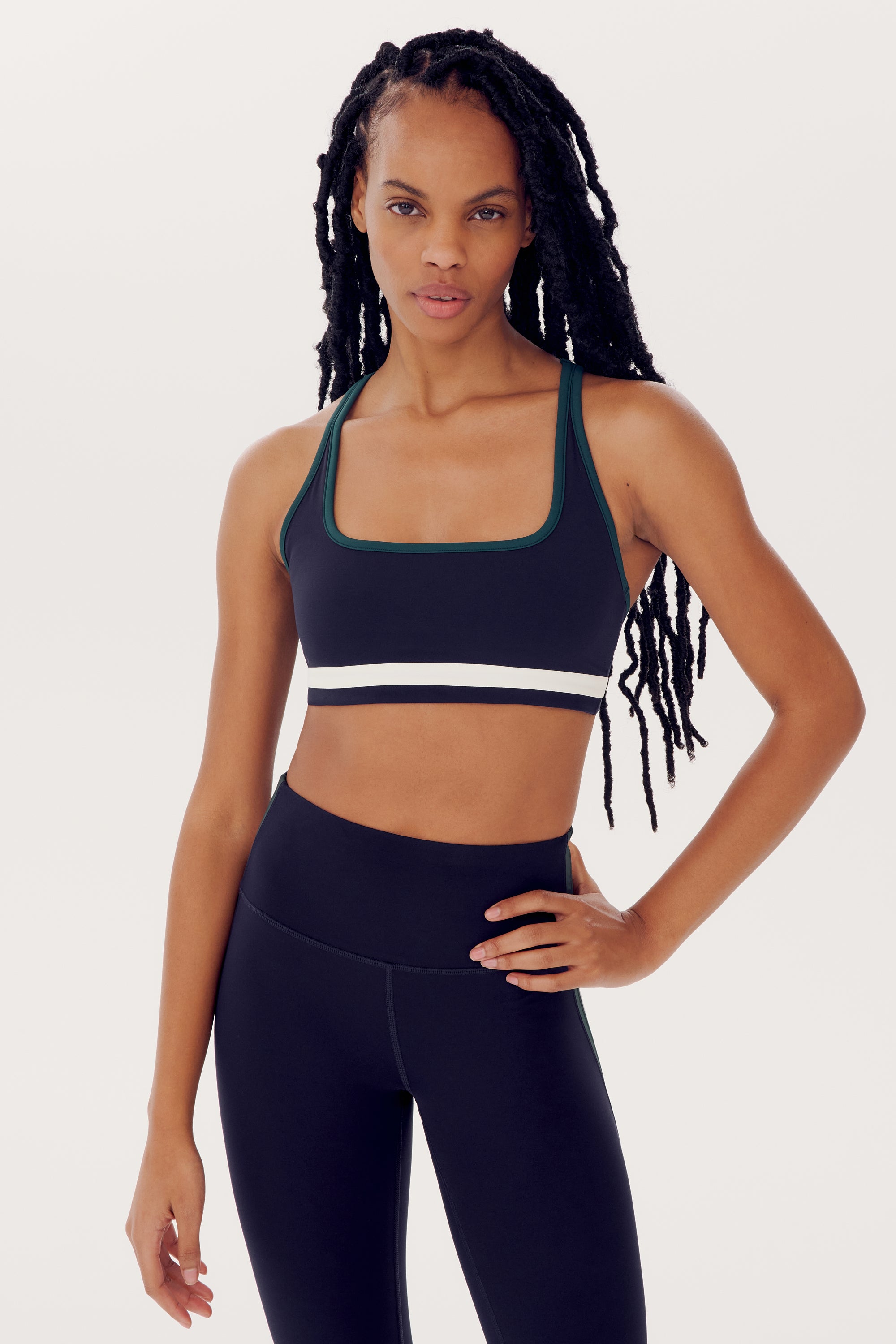 A woman with long braided hair is wearing a Bianca Rigor Bra - Indigo by SPLITS59 and spandex leggings, posing with one hand on her hip against a plain background.