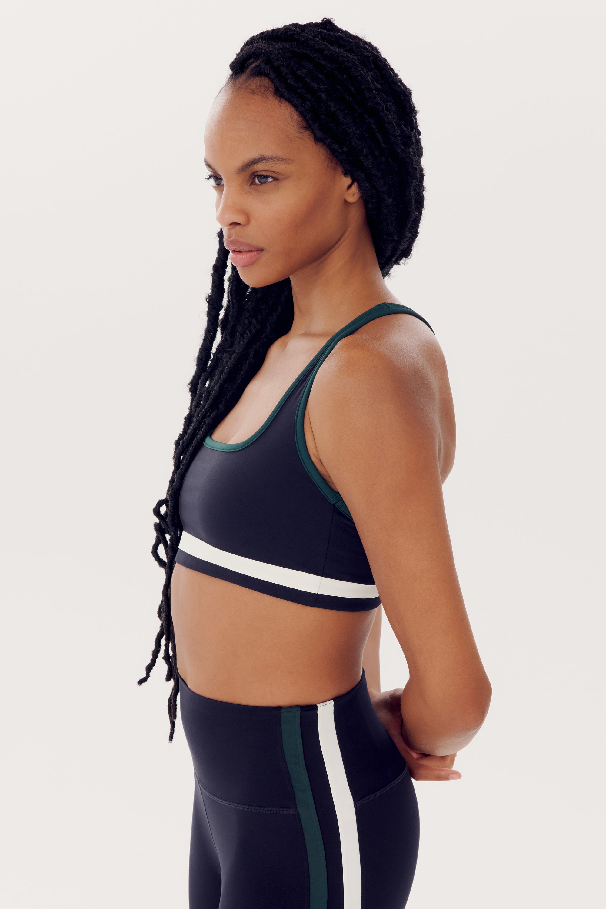 A person with long braided hair, wearing a black Bianca Rigor Bra - Indigo sports bra from SPLITS59 and matching workout leggings made of nylon and spandex, with green and white stripes, stands in profile against a plain background.