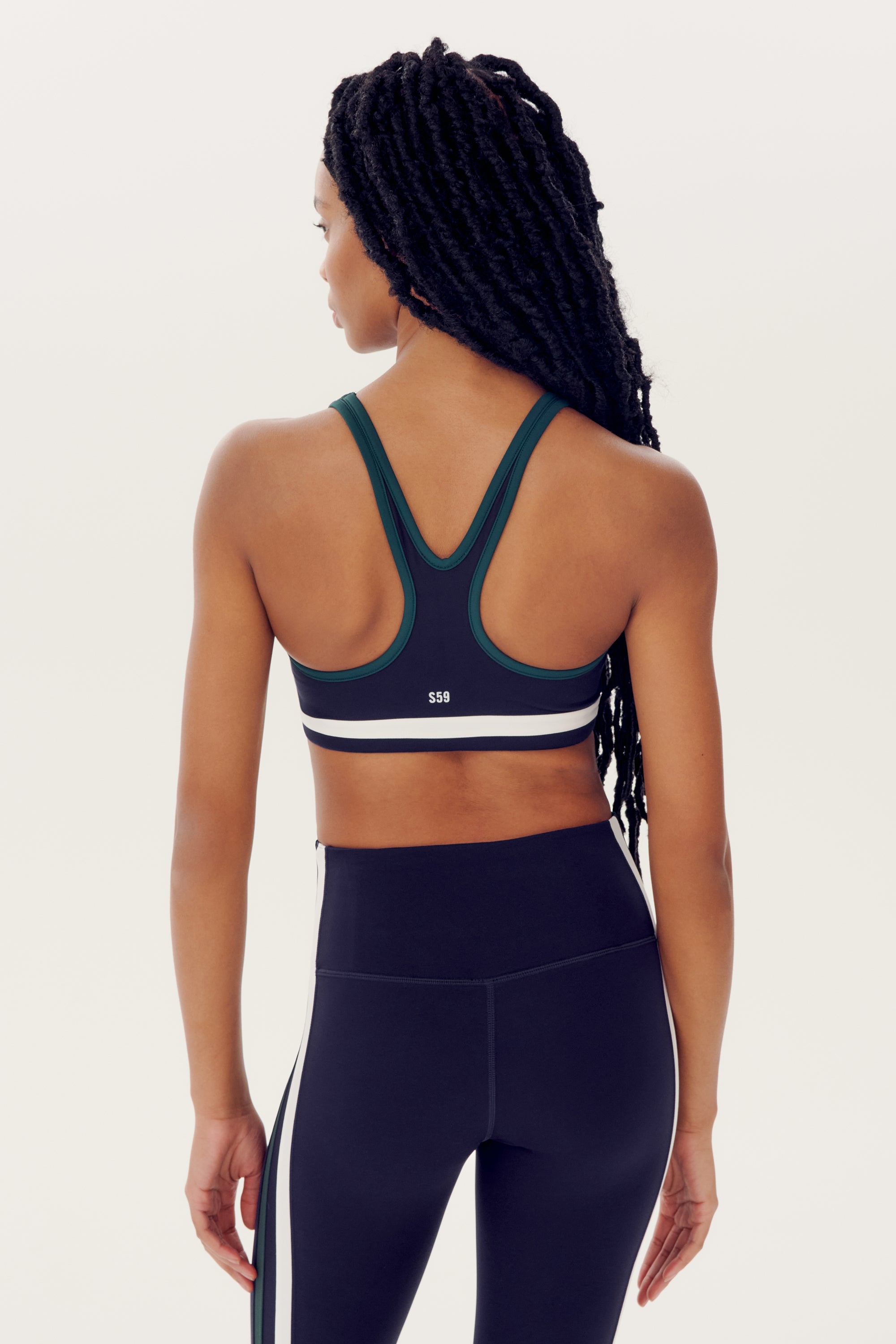 A woman with long braided hair, seen from behind, wears a dark green Bianca Rigor Bra - Indigo by SPLITS59 and black athletic leggings with white side stripes made of nylon and spandex.