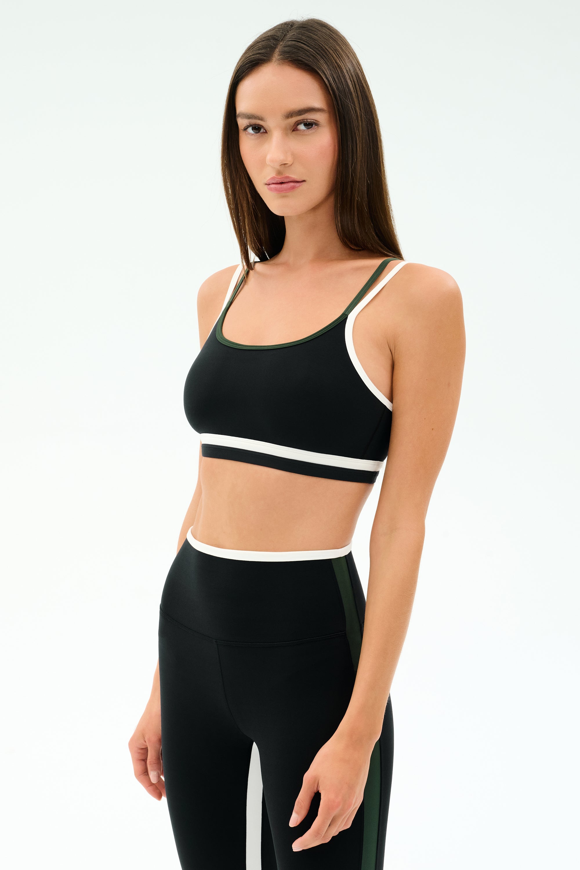 A woman wears the SPLITS59 Eva Rigor Double Strap Bra in Black/Military with matching high-waisted leggings, posing against a plain background—an ideal addition to your workout or yoga bra collection.