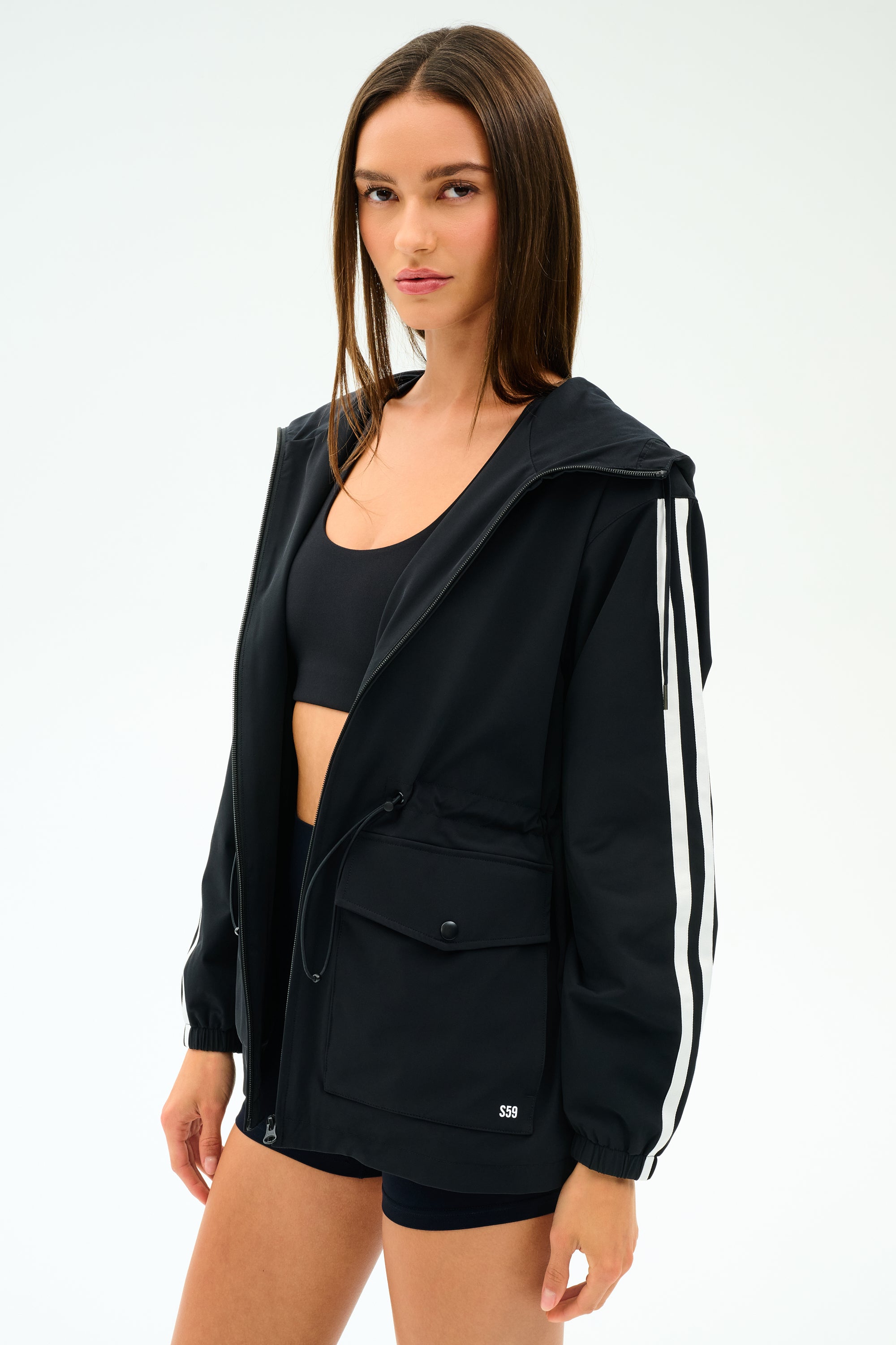 A woman wears the SPLITS59 Reggie Water Repellent Jacket in Black/White over a black sports bra and shorts, standing against a plain white background.