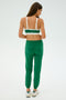 A woman with long brown hair wears a green and white sports bra, Sonja Fleece Sweatpant in Arugula from SPLITS59, and white sneakers. She stands on a plain light background, facing away from the camera.