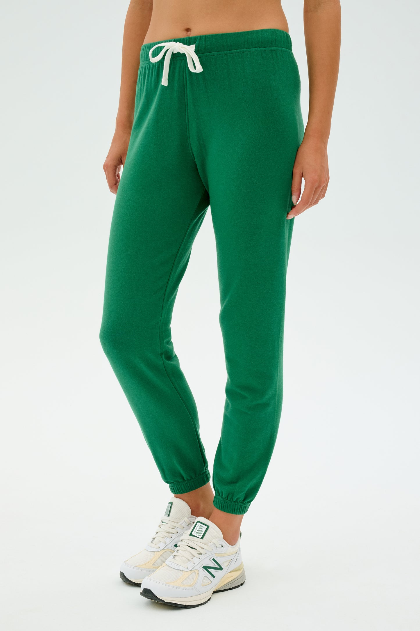 A person dressed in SPLITS59's Sonja Fleece Sweatpant in Arugula and white sneakers stands against a plain background.