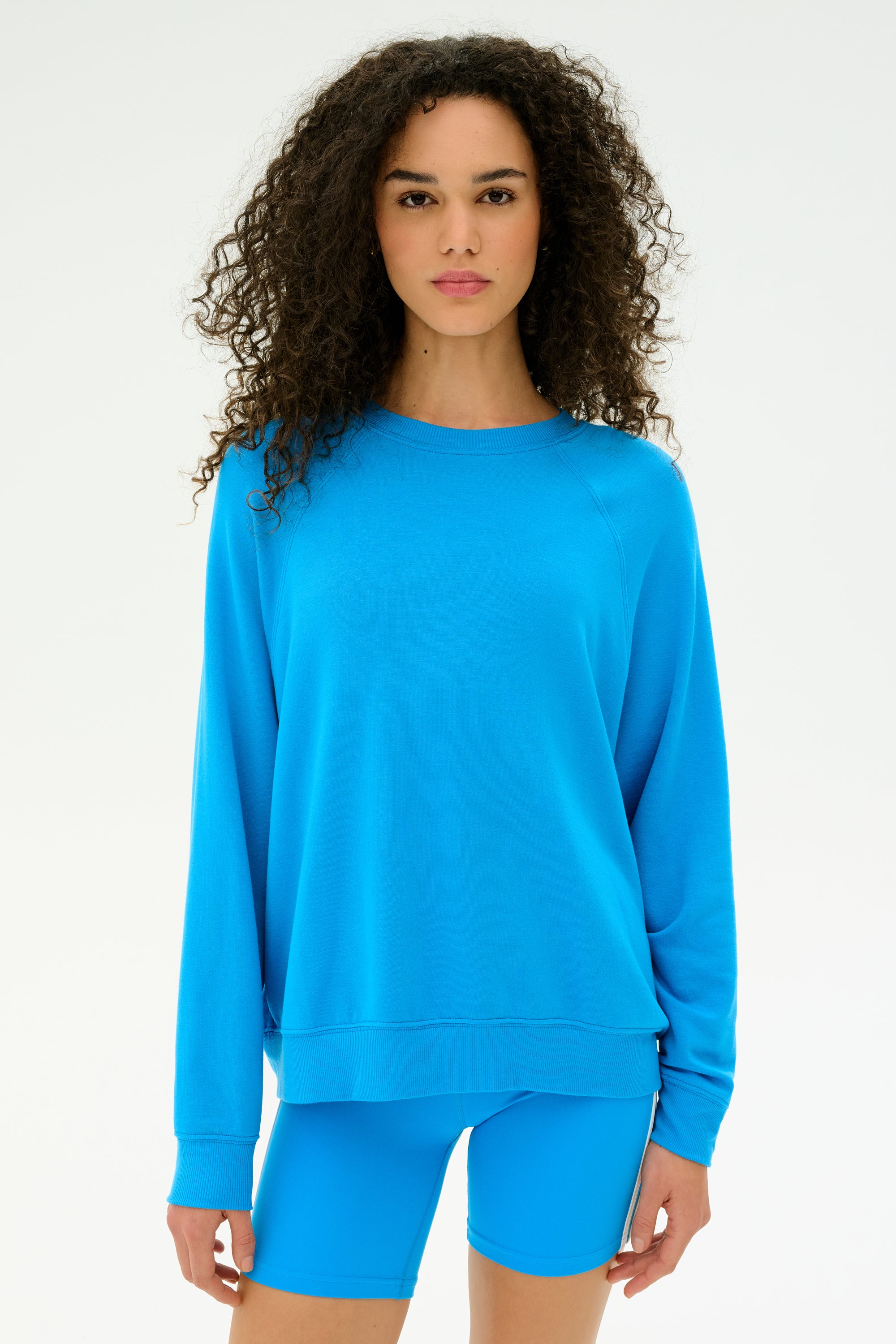 A woman with curly hair wears the SPLITS59 Andie Fleece Sweatshirt in Riviera— a loose, blue crewneck pullover made from soft stretch modal—paired with matching blue shorts, standing against a plain white background.