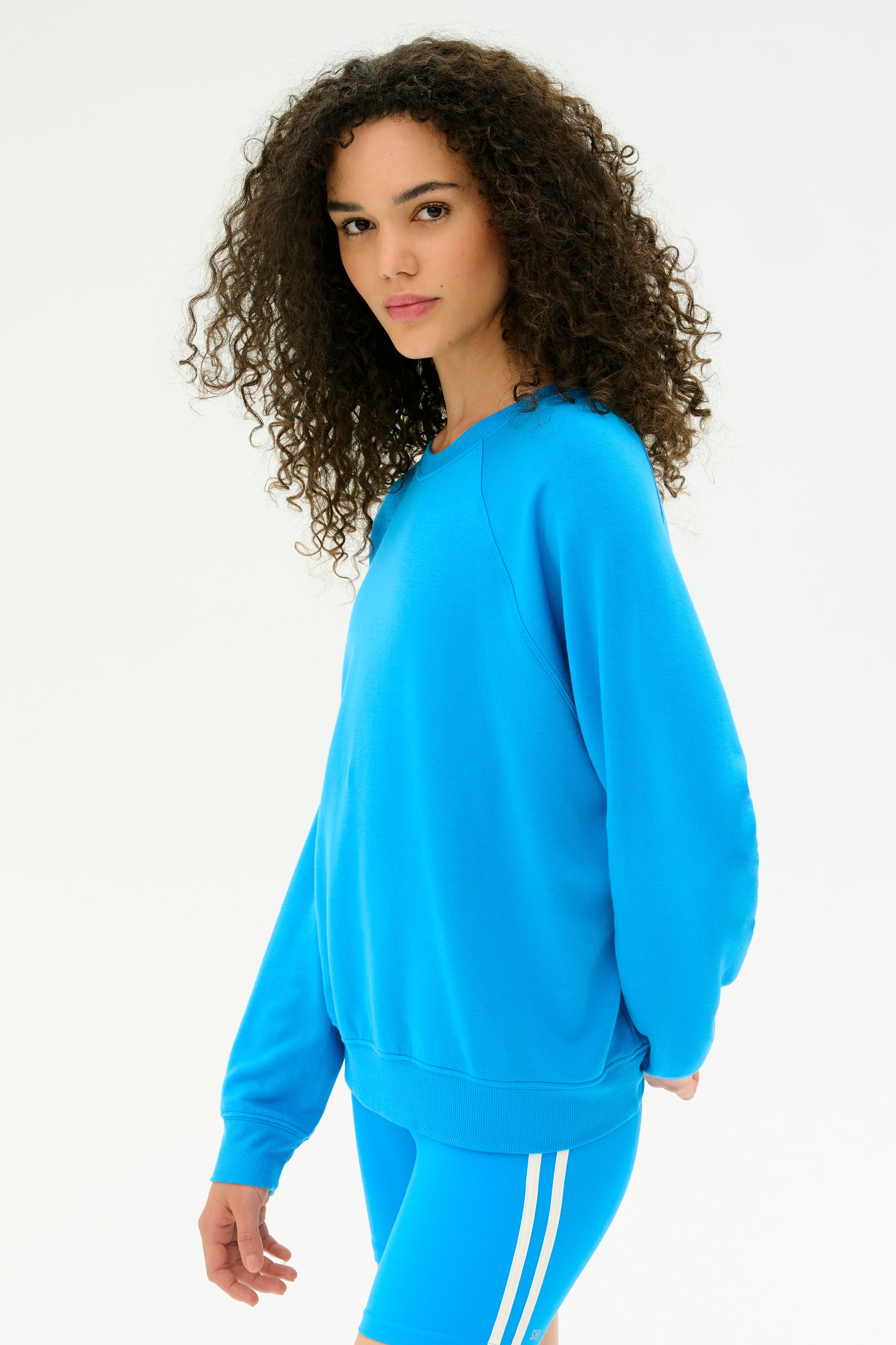 A woman with curly hair wears the SPLITS59 Andie Fleece Sweatshirt in Riviera, paired with blue shorts featuring white stripes. Made from stretch modal and crafted in Los Angeles, she stands against a plain white background.