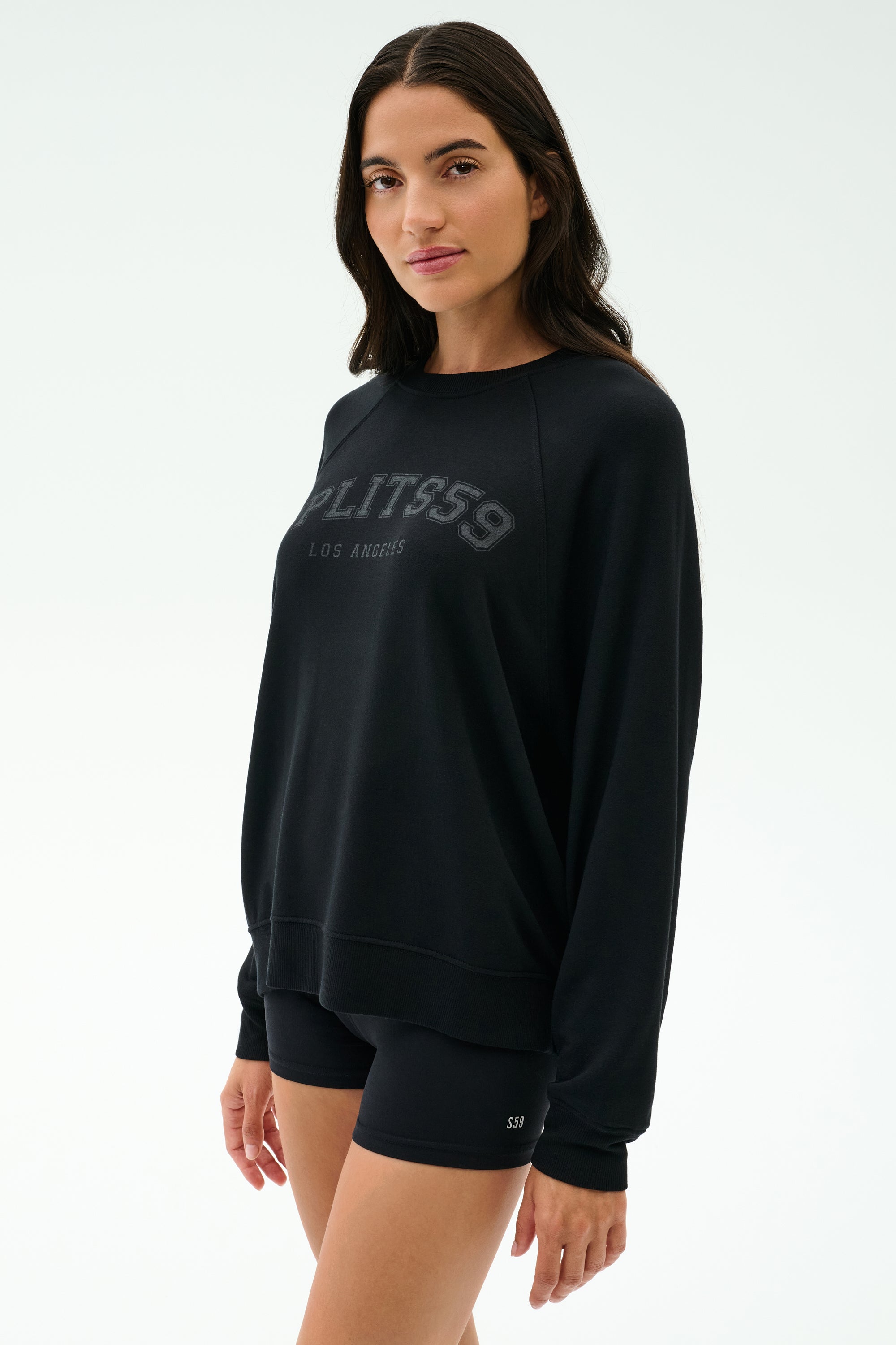A woman wears the SPLITS59 Andie Oversized Fleece Varsity Graphic Sweatshirt in Black/Splits59 Los Angeles and black shorts, made in Los Angeles, standing against a plain light background.