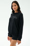 A woman wears the SPLITS59 Andie Oversized Fleece Varsity Graphic Sweatshirt in Black/Splits59 Los Angeles and black shorts, made in Los Angeles, standing against a plain light background.