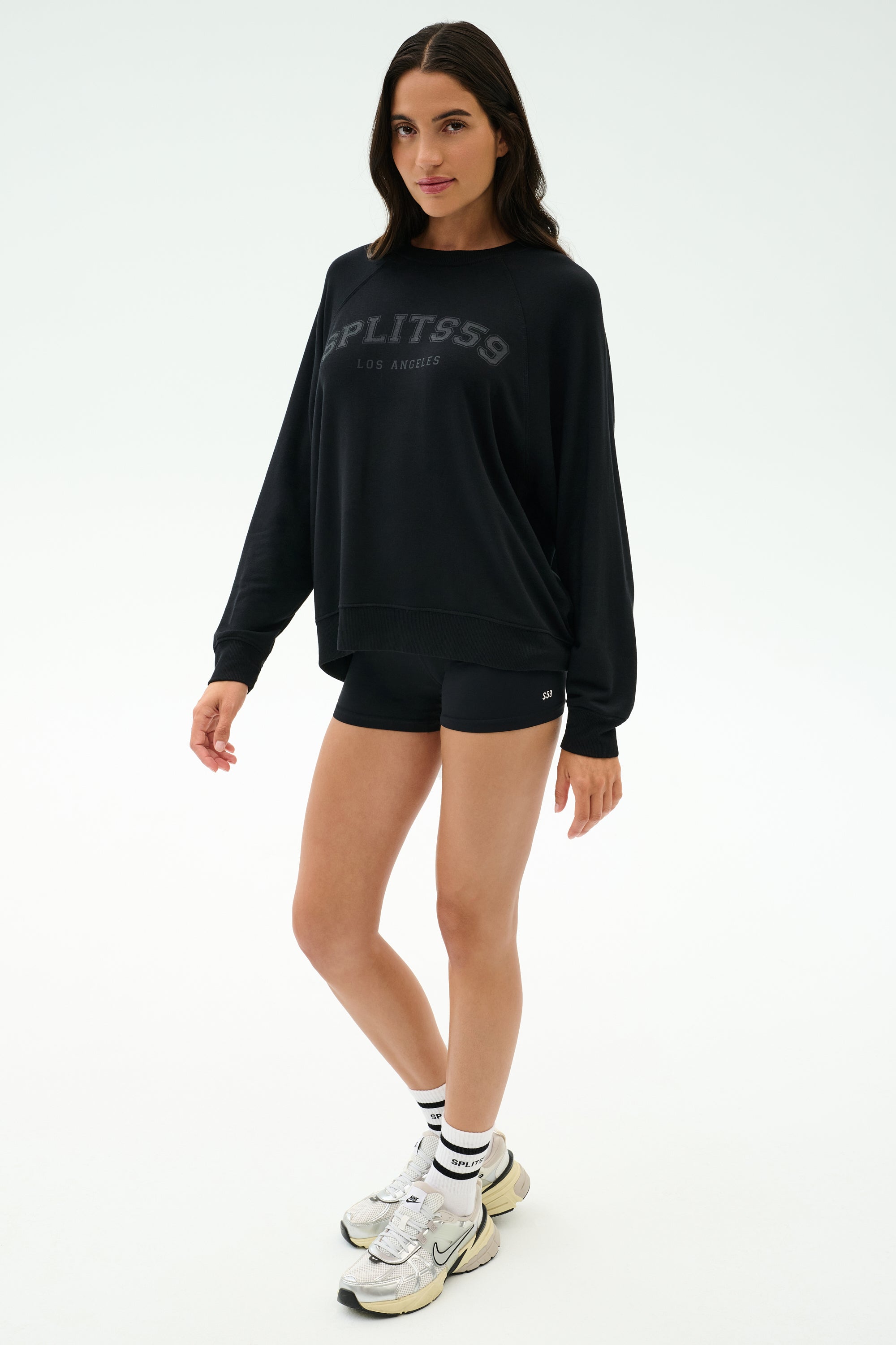 A woman stands in a studio wearing the SPLITS59 Andie Oversized Fleece Varsity Graphic Sweatshirt in black, black shorts, white sneakers, and white socks.